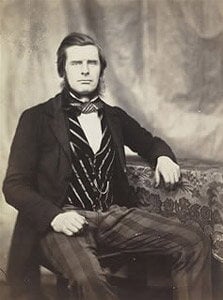 A vintage black and white photograph of a man seated next to a table. He wears a dark suit with a patterned shirt and a bow tie. His hair is styled back, and he gazes directly at the camera. The background is a simple draped curtain.