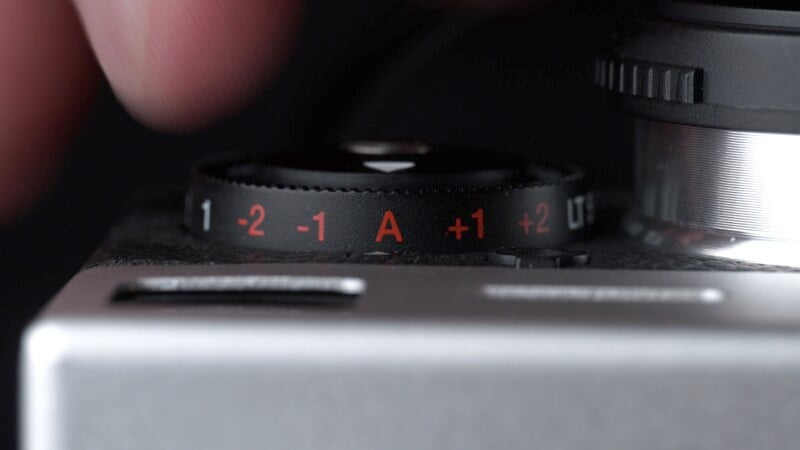 Close-up of a camera dial showing exposure settings: -2, -1, A, +1, +2. A hand is visible adjusting the dial, and part of the camera body is in the foreground.