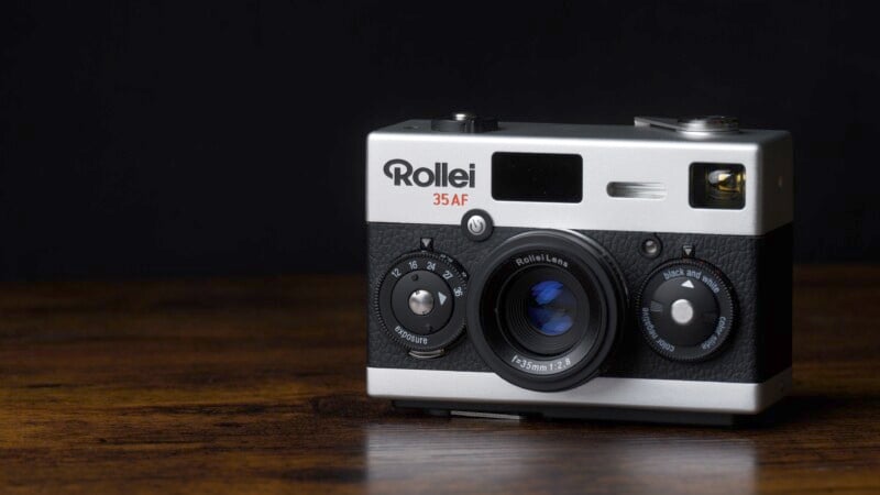 A vintage Rollei 35 AF camera sits on a wooden surface against a black background. The camera features a metallic silver body, black lens, and dials, showcasing a classic design.