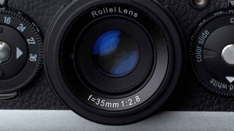 A close-up of a camera lens labeled "Rollei Lens f=35mm 1:2.8." The lens is surrounded by a textured black surface with dials on either side, indicating a vintage or classic camera design.