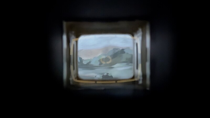 A view through a small, rectangular opening showing a blurry landscape painting with muted colors, depicting mountains, sky, and water. The scene is framed by a dark, tunnel-like border, suggesting a distant, dreamlike perspective.