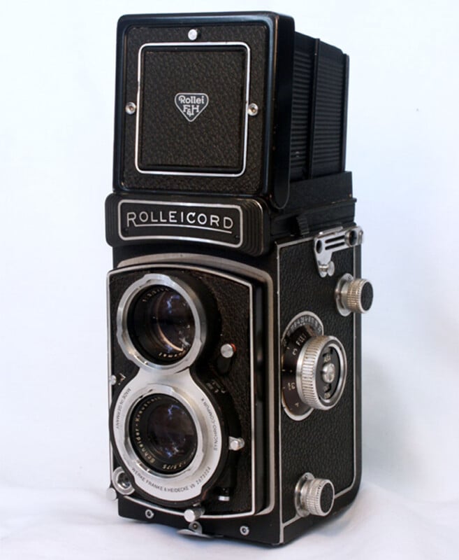 A vintage Rolleicord twin-lens reflex camera with black and silver detailing. It features two lenses stacked vertically on the front, surrounded by knobs and dials, against a plain white background.