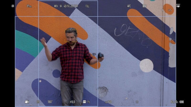 A person in a red and black plaid shirt poses playfully against a colorful, abstract mural with blue, orange, and white shapes. They hold a camera and appear within the frame of a camera screen, with settings displayed.