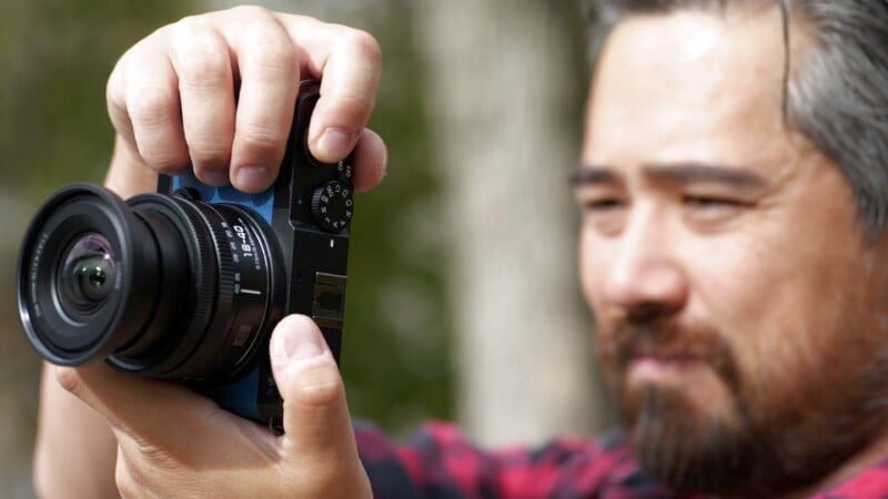 A person with a beard, wearing a red plaid shirt, is holding a camera up to their eye, seemingly about to take a photo. The background is blurred, suggesting an outdoor setting.