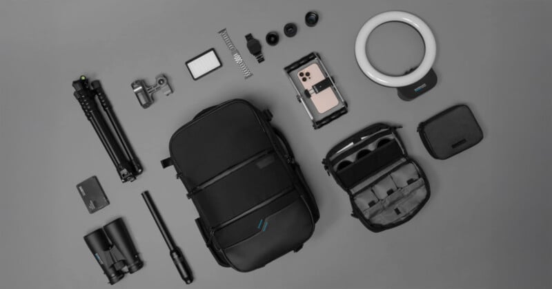 A collection of photography gear arranged on a gray background. Includes a black backpack, binoculars, camera tripod, lens kit, smartphone gimbal, LED ring light, small carrying case, and various camera accessories.