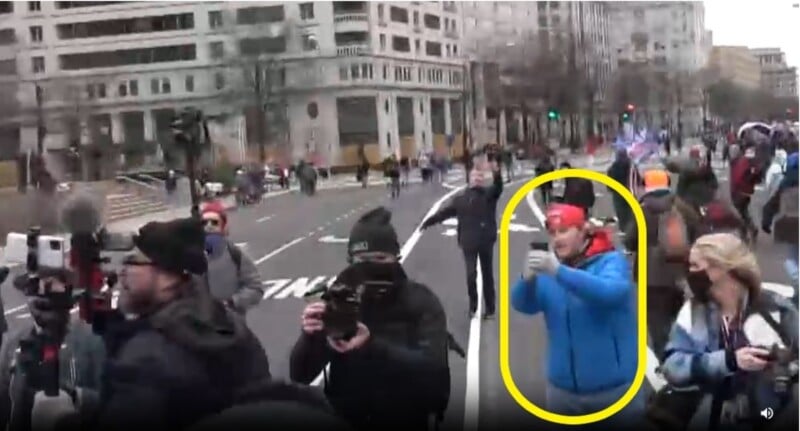 A group of people on a city street, some are holding cameras. Buildings line the background. One person in a blue jacket and red cap, inside a yellow oval, is taking a photo or recording. It's a cloudy day.