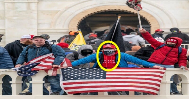 A group of people stand on a building's steps, some holding American flags. The central figure is wearing red and holding a flagpole. The scene appears chaotic, with people dressed for cold weather.