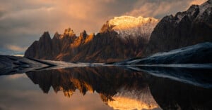 A serene landscape of jagged mountains reflecting in a calm lake. The peaks are illuminated by a warm, golden sunset, contrasting with the dark, rocky foreground and slightly cloudy sky.