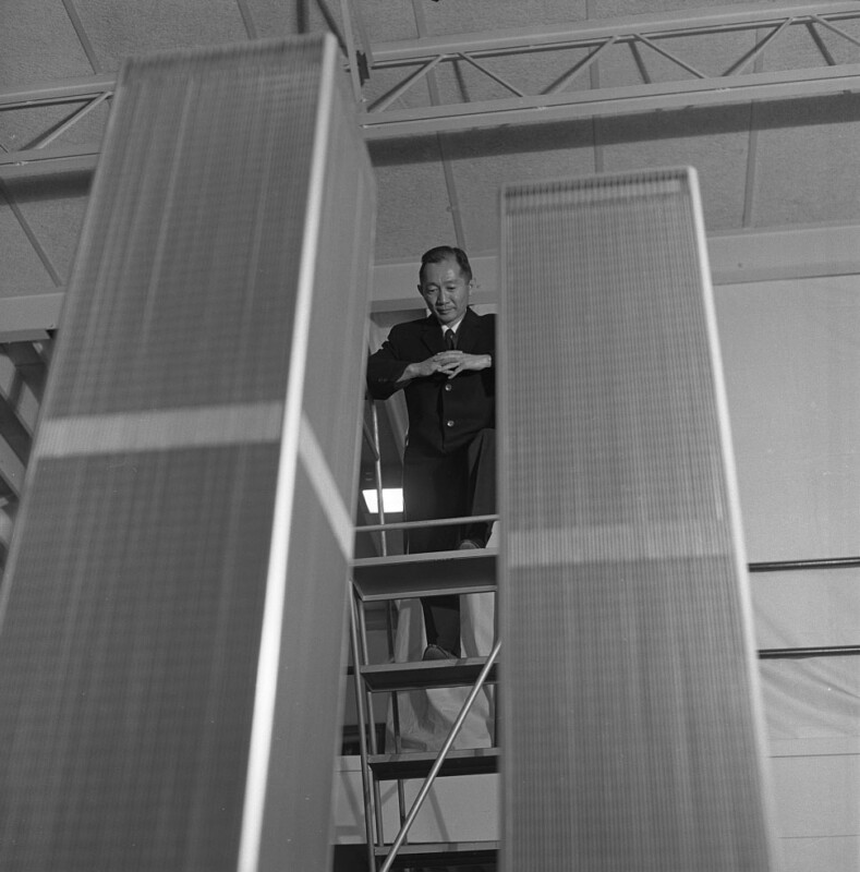 A man in a suit stands on a platform, looking down at two tall, rectangular architectural models in the foreground. He seems to be observing the models thoughtfully with his hands clasped. The background shows structural elements of the building.