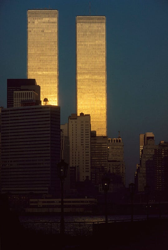 The image shows the silhouette of the New York City skyline at sunset, prominently featuring the Twin Towers of the World Trade Center bathed in golden light. Several other skyscrapers and buildings are visible in front of and around the towers.