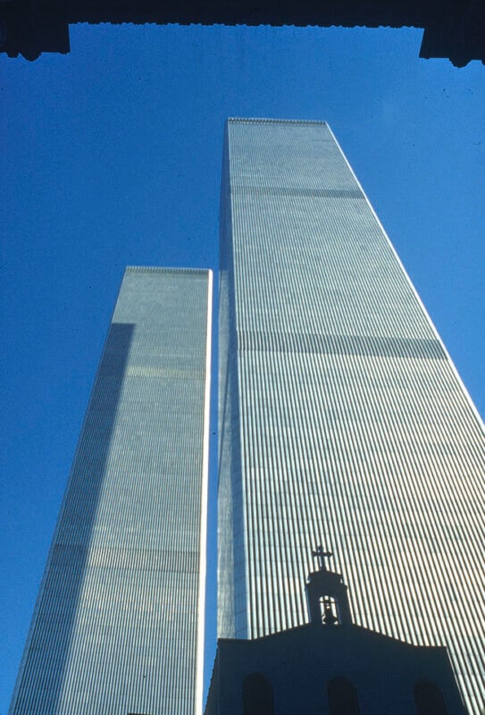 A photograph of the Twin Towers of the World Trade Center in New York City. The towers are seen from a low angle, reaching up into a clear blue sky. The top part of a building with a small dome and cross is visible at the bottom of the image.