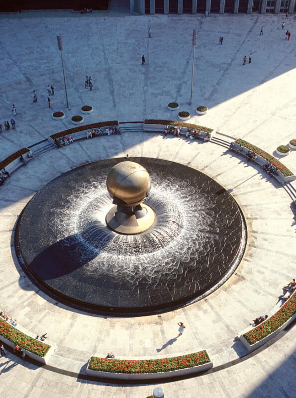 An aerial view of a large, circular fountain with a metal sphere at its center, surrounded by flowing water. The fountain is situated in a spacious, open plaza with a few people walking and sitting nearby. The area is bordered by flower beds and benches.