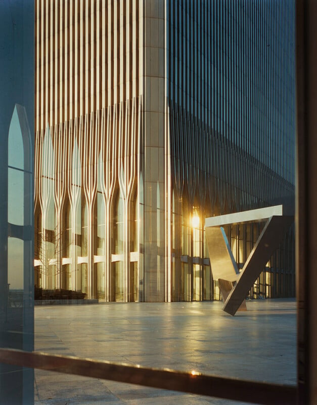 A tall, modern building with vertical lines and a sunlit exterior stands beside an open plaza. The lower part of the building features elegant arch-like designs, and a large, angular metallic sculpture is positioned nearby. The image captures the scene at sunrise or sunset.