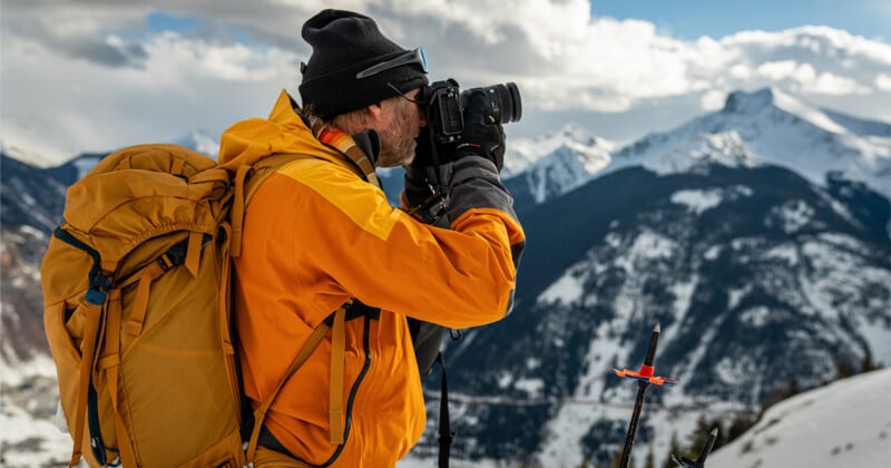 A person in an orange jacket and dark beanie uses a camera in a snowy mountain landscape. They have a large backpack and are surrounded by snow-covered peaks under a partly cloudy sky.