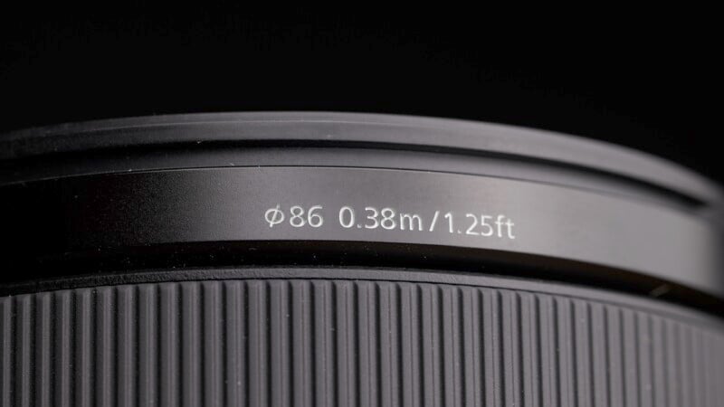 Close-up of a camera lens with etched specifications: ϕ86 0.38m/1.25ft. The lens features a textured focus ring, and the lighting highlights the details against a dark background.