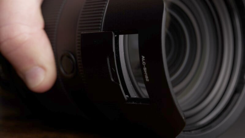 Close-up of a person holding a large camera lens with a lens hood attached. The focus is on the intricate details of the lens elements visible through the glass, emphasizing the precision and craftsmanship of the camera equipment.