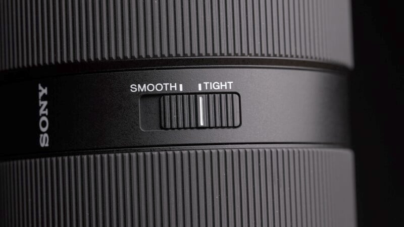 Close-up of a camera lens showing a switch labeled "Smooth" and "Tight." The switch is positioned in the middle. The lens is black with textured grip sections, and part of the brand name is visible on the side.