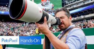 A photographer with a large telephoto lens takes a picture at a crowded outdoor sports event. The stadium is filled with spectators, and a large screen is visible in the background. The image has a "PetaPixel Hands-On" text overlay.