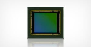 Square image sensor with a gradient from green to blue in the center, surrounded by a black border with gold contacts. The sensor is set against a light background, and its reflection is visible below.