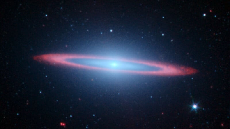 A bright, elliptical galaxy resembling a glowing eye against a black backdrop of space speckled with white and red stars. The galaxy features a vibrant blue center and glowing red edges, creating a halo effect.