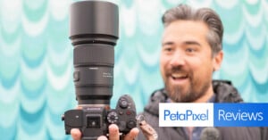 A man holds a Sony camera with a large Tamron lens, with a blurred teardrop pattern background. There is a "PetaPixel Reviews" logo in the corner. He appears to be engaged with the camera.