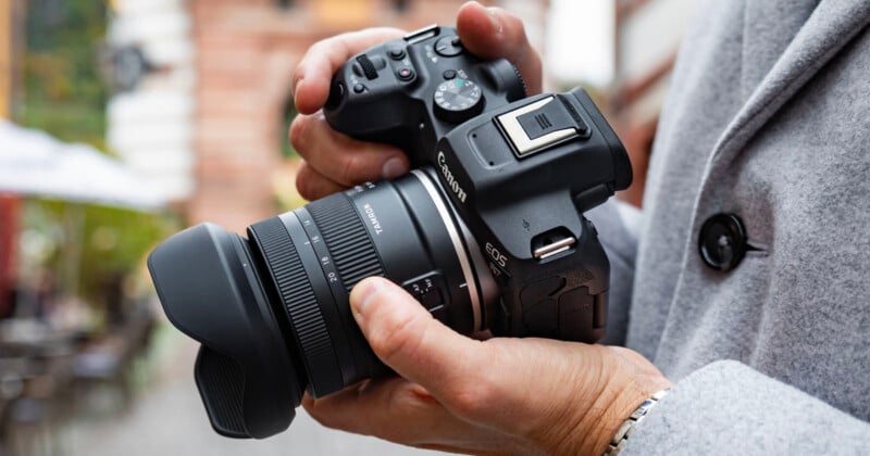 Person holding a Canon EOS camera with both hands, focusing on the camera lens. Background is slightly blurred, indicating an outdoor setting. Person is wearing a light gray coat.