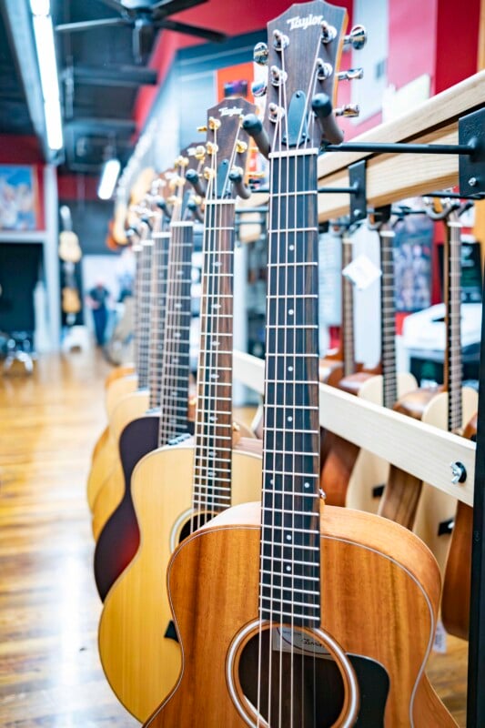 A line of acoustic guitars displayed vertically in a music store, each slightly different in wood finish. The guitars are positioned on stands, and the store's interior, with wooden flooring and a blue ceiling, is visible in the background.