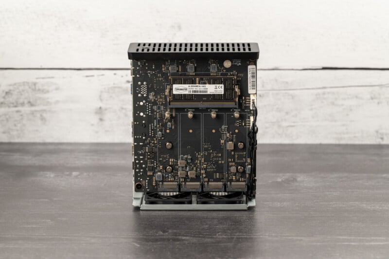 A vertically standing computer motherboard with visible circuits, components, and ports. Two RAM sticks are installed, along with various capacitors and chips. The background features a rough, white wooden surface, and the setup is placed on a dark tabletop.