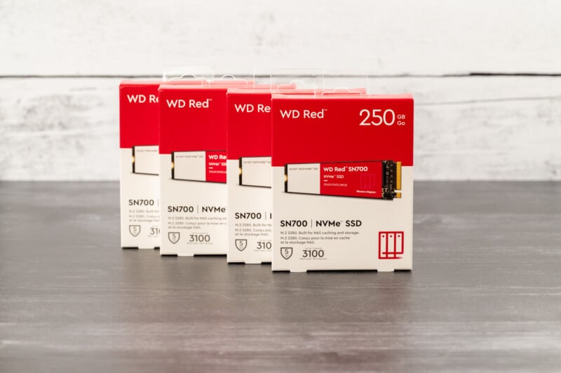 Five boxes of WD Red SN700 NVMe SSDs, stacked in a row, are displayed on a dark surface. The boxes are predominantly red and white, each labeled with "250 GB" and featuring an image of the SSD. The background is a light-colored wooden texture.