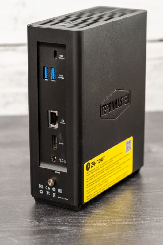 A TerraMaster network-attached storage (NAS) device is shown, standing upright on a dark surface. It has multiple ports, including USB, Ethernet, HDMI, and a reset button. The device is black with a yellow sticker providing information on the bottom section.