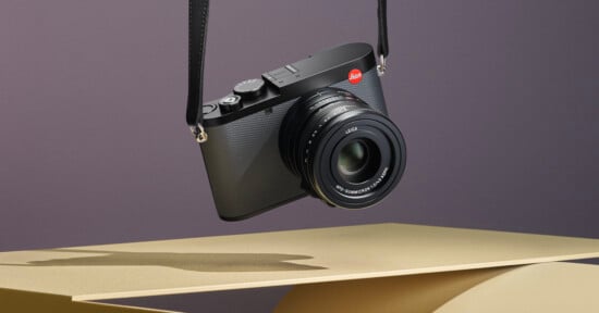 A black Leica camera with a textured body is suspended by its strap against a neutral background. The camera features a large lens with visible markings. It is positioned above abstract, geometric shapes in shades of beige and gold.