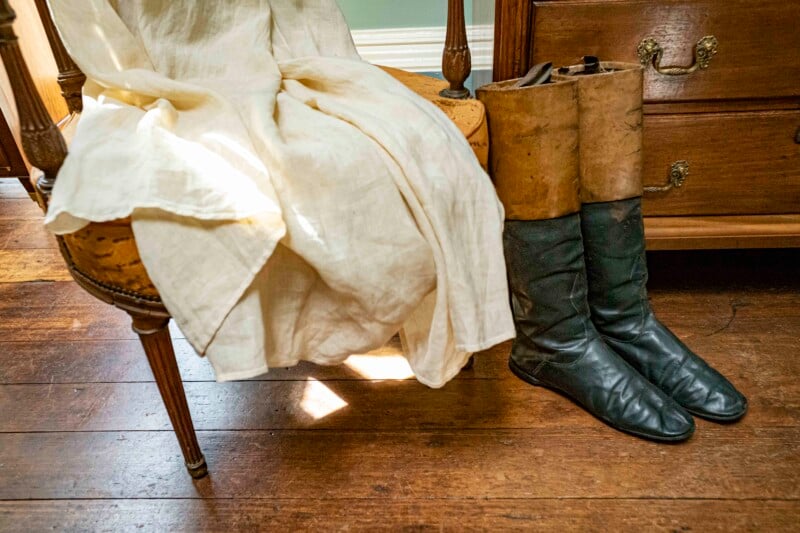 A rustic room with a wooden floor features an upholstered chair draped with a cream-colored fabric. Beside the chair, worn-out tall leather boots lean against a wooden dresser with ornate handles. Sunlight softly illuminates the scene.