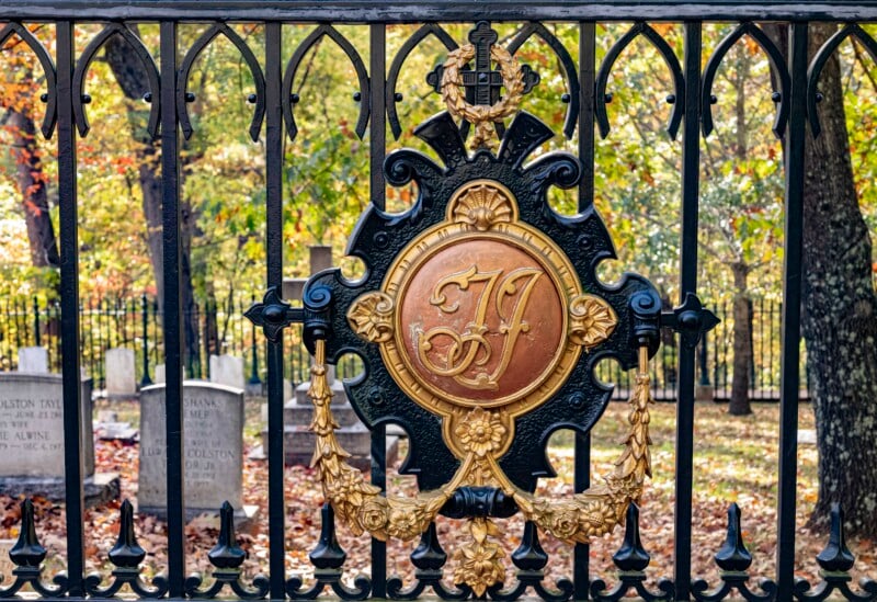 An ornate wrought iron fence in a cemetery, featuring a decorative emblem with the initials "JF" in gold. In the background, tombstones are visible among autumn trees with colorful leaves.