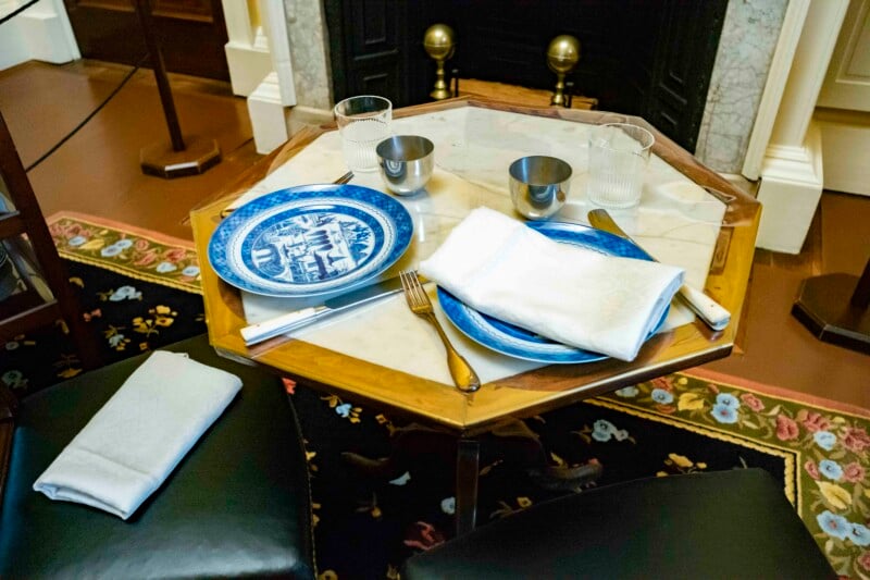 A small dining table with a marble top set for two, featuring blue and white china plates, silver cutlery, two glass tumblers, and two metal cups. A floral-patterned carpet and part of a fireplace are visible in the background.