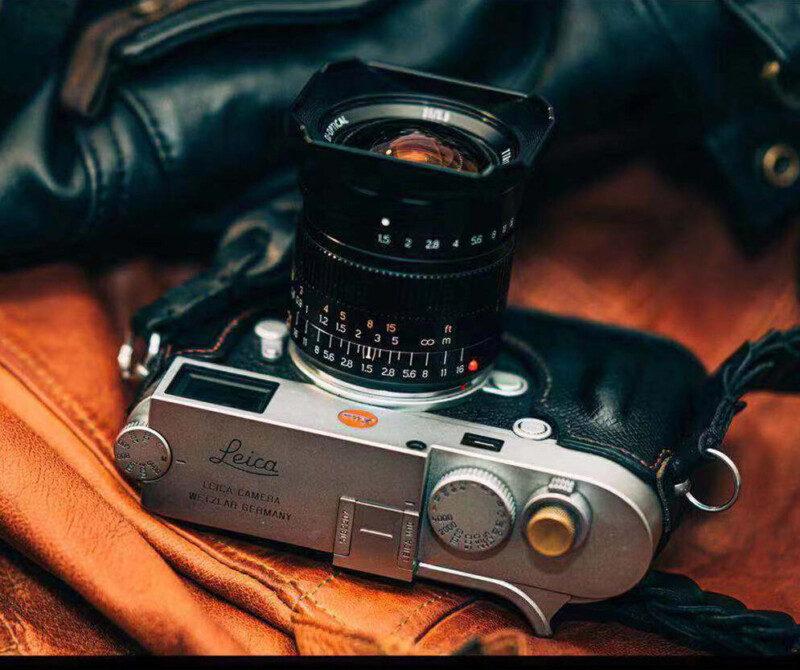 A vintage Leica camera with a wide-angle lens rests on a brown leather surface, surrounded by a soft black cloth. The camera features a silver body with various dials and engraved branding.