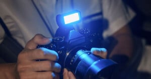 A person is holding a black DSLR camera with an attached external flash that is emitting blue light. The camera lens and part of the person's hands are in focus, while the background is blurred.