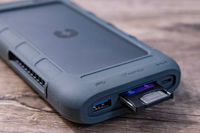 A close-up image of a rugged external hard drive with a grey protective casing. The drive has ports and slots for USB and other connectivity options, with one connector partially inserted. The device is placed on a wooden surface.