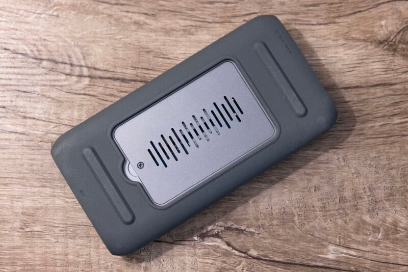 A rectangular, dark grey portable speaker sits on a wooden surface. The speaker has a metal grille with vertical slits in the middle, surrounded by a rugged outer casing. The design is minimalistic and sturdy, suitable for outdoor use.