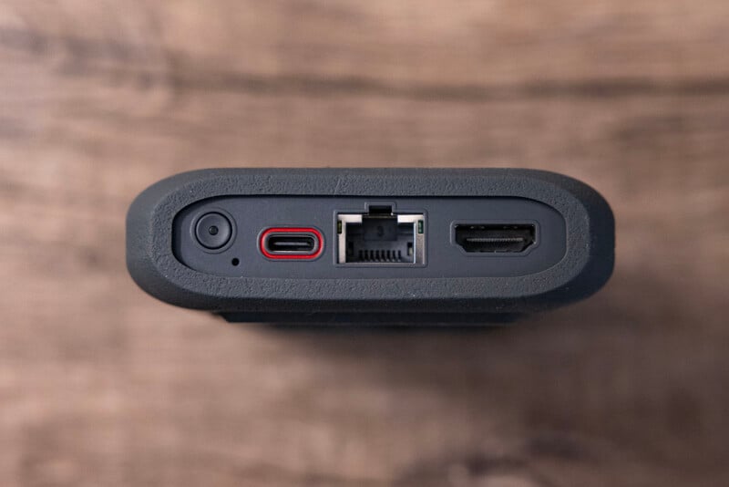 Close-up view of an electronic device's ports: a small circular power port, a USB-C port with a red ring, an Ethernet port, and an HDMI port. The background is a wooden surface.