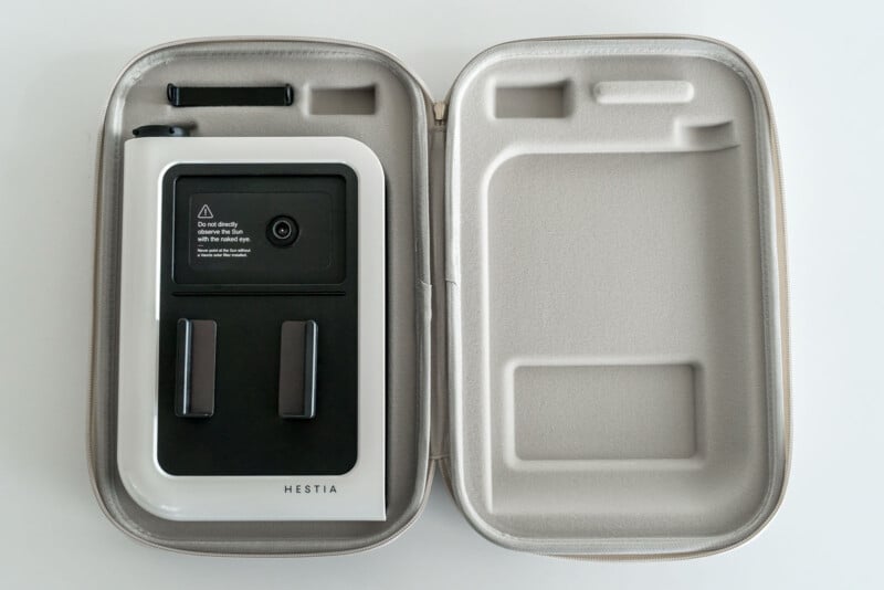 An open carrying case displaying a white and black electronic device labeled "HESTIA" on one side. The device is secured in a fitted compartment alongside an empty section presumably for accessories. The interior of the case is gray with molded fittings.