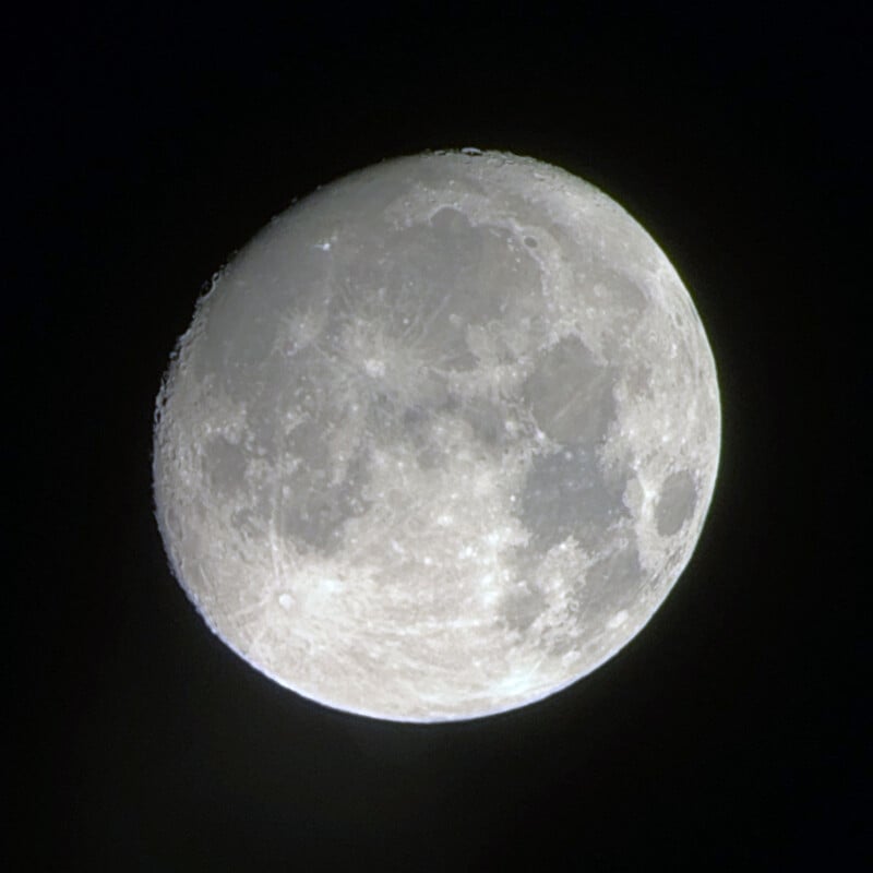 A detailed view of the moon, showing its cratered and textured surface against a dark sky. The moon is predominantly illuminated, showcasing its bright, reflective regions and several patches of darker areas.