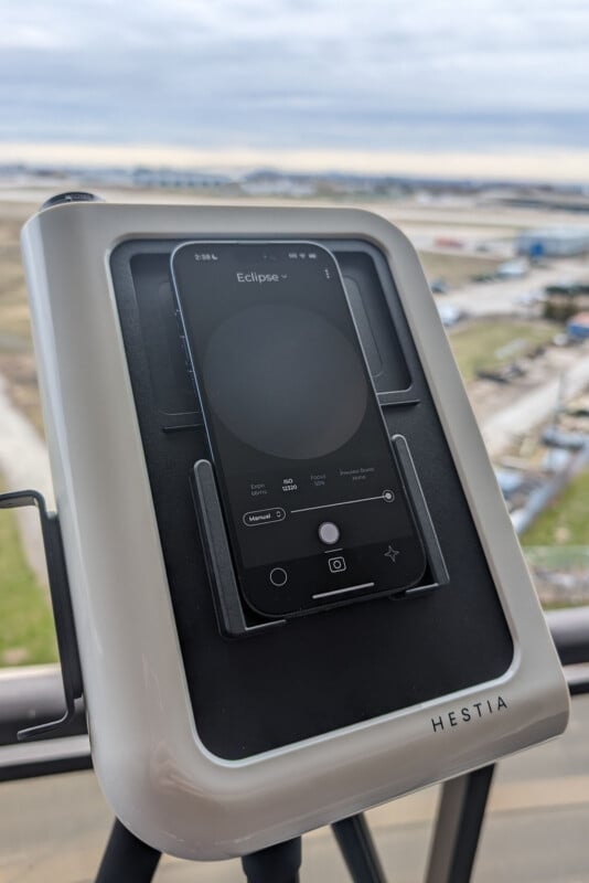 A smartphone is mounted on a Hestia device, which is standing on a tripod. The device is outdoors, with a blurred view of a landscape and cloudy sky in the background. The phone screen displays an app named Eclipse.