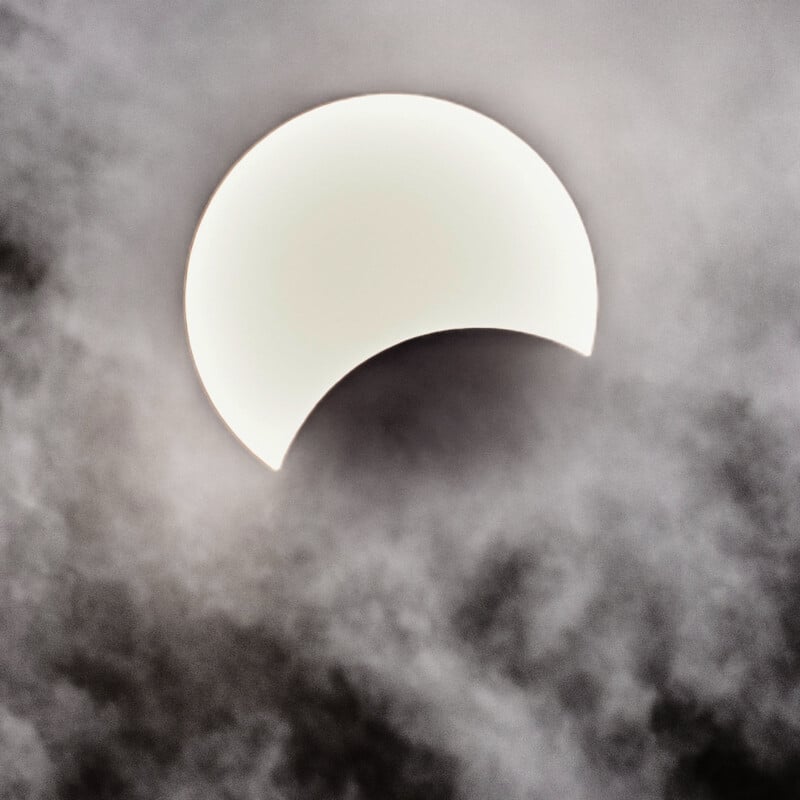 A partially eclipsed sun is visible behind a layer of clouds, creating a dramatic effect. The moon has covered a portion of the sun, forming a crescent shape with a bright, glowing edge. The surrounding sky is cloudy and gray.