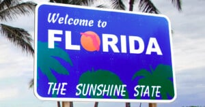 A road sign reads "Welcome to Florida, The Sunshine State" with a stylized orange in place of the "o" in Florida. Palm tree silhouettes are in the background, set against a cloudy sky.