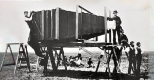 A black and white historical photograph shows several men standing around and on a giant camera, mounted on a large wooden platform. Some men are posing on ladders and support beams, while others sit on the ground in the background.