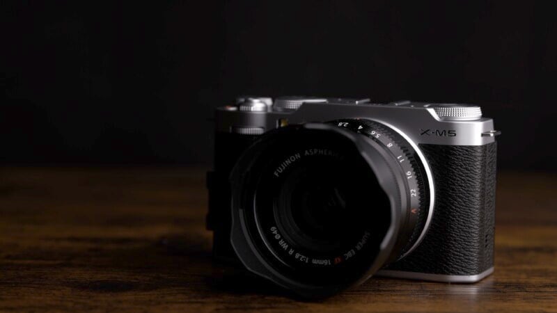 A vintage-style, silver and black camera with a lens marked with various numbers is placed on a dark wooden surface. The lighting is low and focused on the camera, casting soft shadows against a dark background.