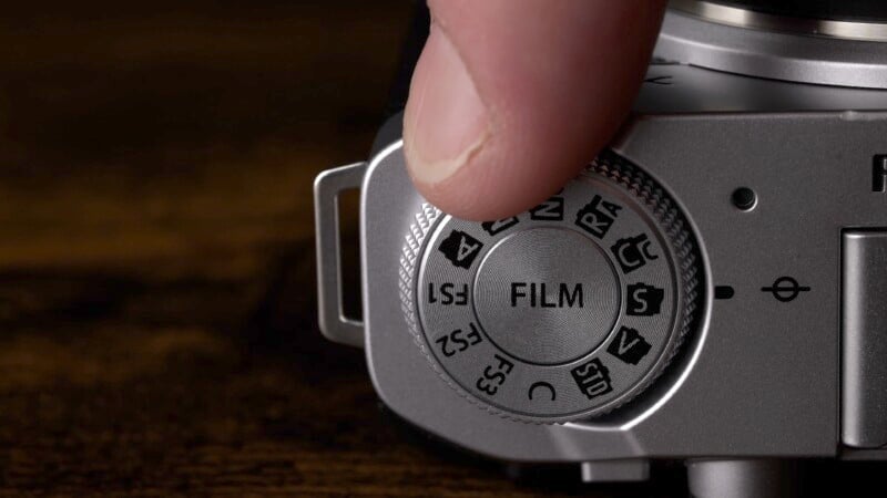 A close-up of a person's finger adjusting a camera dial set to "FILM." The dial includes various settings like P, A, S, M, and different scene modes. The background is a dark wooden surface.