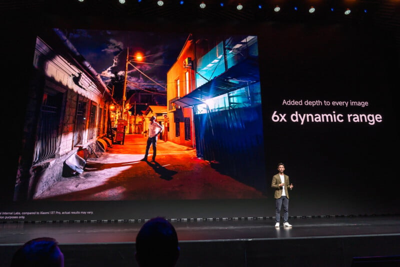 A speaker on a stage gives a presentation, standing in front of a large screen displaying an image of a person standing on a dimly lit street at night. The screen text reads: "Added depth to every image, 6x dynamic range.