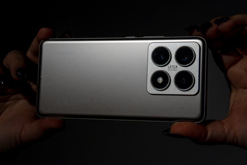 A pair of hands with dark nail polish holds a silver smartphone horizontally. The back of the phone features three prominent camera lenses and a smaller lens arranged in a square module, with "Leica" inscribed on it. The brand name is subtly visible on the back.