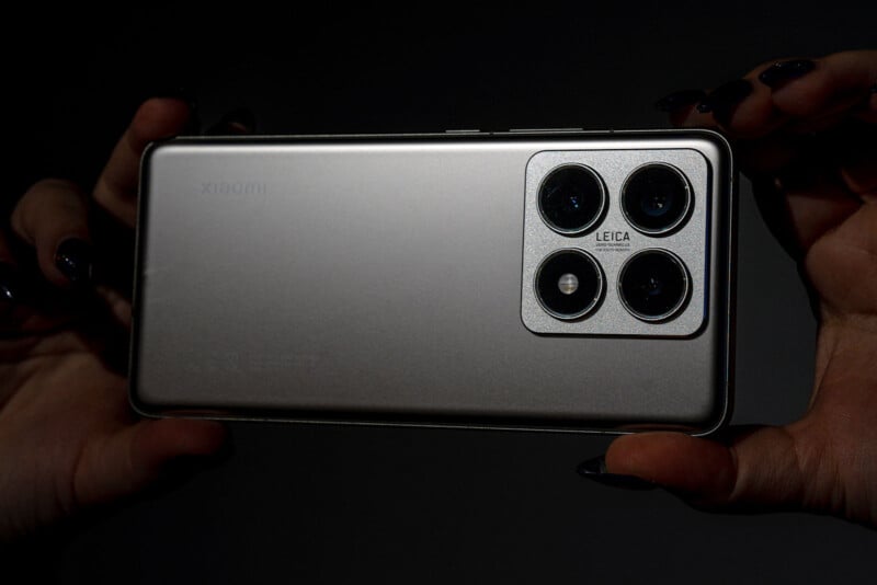 Close-up of a person holding a sleek silver smartphone horizontally. The phone features a triple camera system with a prominent rectangular module in the upper corner. The brand name "Leica" is visible near the camera lenses.